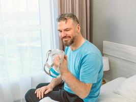 gelukkig uitgeslapen man met chronische ademhalingsproblemen na het gebruik van een cpap-machine zittend op het bed in de slaapkamer. gezondheidszorg, cpap, obstructieve slaapapneu-therapie, snurkconcept foto