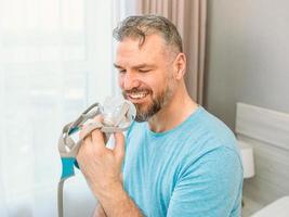 volwassen gelukkige man met chronische ademhalingsproblemen overweegt zittend op het bed in de slaapkamer met cpap-machine. gezondheidszorg, cpap, snurkconcept foto