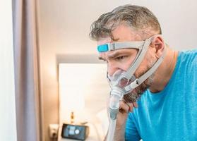 ongelukkige geschokte man met chronische ademhalingsproblemen verrast door het gebruik van een cpap-machine die op het bed in de slaapkamer zit. gezondheidszorg, cpap, obstructieve slaapapneu-therapie, snurkconcept foto