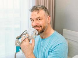 volwassen gelukkige man met chronische ademhalingsproblemen overweegt het gebruik van cpap-machine zittend op het bed in de slaapkamer. gezondheidszorg, cpap, snurkconcept foto