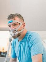 ongelukkige geschokte man met chronische ademhalingsproblemen verrast door het gebruik van een cpap-machine die op het bed in de slaapkamer zit. gezondheidszorg, cpap, obstructieve slaapapneu-therapie, snurkconcept foto