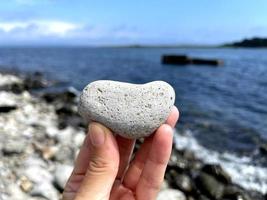 hartvorm steen in meisjes hand tegen de achtergrond van het strand. zomer zonnige dag. liefde, huwelijk en valentijnsdag concept. mooie en interessante stenen vinden. strandvakantie foto