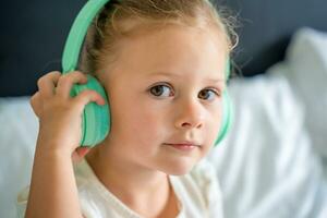 schattig weinig meisje genieten van muziek- gebruik makend van groen kinderen hoofdtelefoons in huis bed. hoog kwaliteit foto