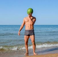 een sportief Mens is staand Aan de strand en Holding een geheel watermeloen. een rijp watermeloen in de handen van een Mens foto