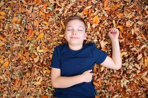 portret van een 9 jaar oud blond jongen in een herfst park. een knap jongen leugens in oranje gedaald bladeren, looks Bij de camera en glimlacht. zonnig dag in de park. foto