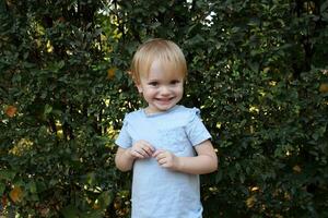 portret van een 2 jaar oud blond meisje Aan een achtergrond van bladeren. Kaukasisch todler met groot ogen looks Bij de camera en glimlacht. foto