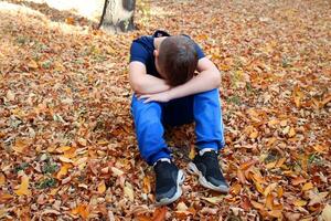 portret van een 9 jaar oud jongen in de herfst park. een jongen in een blauw t-shirt zit in oranje gedaald bladeren en zet zijn hoofd Aan zijn knieën. foto