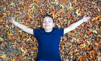 portret van een 9 jaar oud blond jongen in een herfst park. een knap jongen leugens in oranje gedaald bladeren, looks Bij de camera en glimlacht. zonnig dag in de park. foto