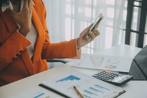 glimlachen jong Aziatisch zakenvrouw werken Aan een laptop computer Bij haar bureau in een helder modern huis kantoor, aan het doen berekenen kosten financieel verslag doen van financiën maken aantekeningen Aan papier diagram gegevens document. foto