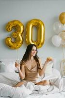 gelukkig vrouw houdt een feestelijk taart in haar handen terwijl zittend Aan de bed in de slaapkamer versierd voor de vakantie en een glas van Champagne in eer van haar verjaardag foto