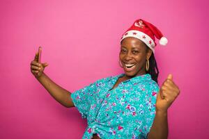 overenthousiast zwart Afrikaanse vrouw millennium vieren met haar slim telefoon na ontvangen mooi zo nieuws. vervelend Kerstmis hoed staand achter een roze studio muur. Kerstmis concept foto