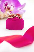 goud bruiloft ringen Aan roze fluweel hart Aan geschenk doos met orchideeën Aan wit achtergrond. huwelijk, voorstel, Valentijnsdag dag. verticaal. kopiëren ruimte foto