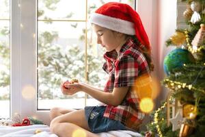 meisje in een de kerstman hoed zit Aan de vensterbank van een huis in de buurt de Kerstmis boom en Holding glas ballen in zijn handen. kind is hebben plezier, gelooft in een wonder en aan het wachten voor Kerstmis en nieuw jaar foto