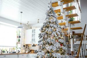 feestelijk Kerstmis decor in wit keuken, modern dorp interieur met een besneeuwd Kerstmis boom en fee lichten. nieuw jaar, Kerstmis stemming, knus huis, metaal modulair trappenhuis. de algemeen plan foto