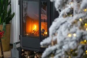 Metall zwart fornuis, brandend haard haard in wit feestelijk interieur van huis is versierd voor Kerstmis en nieuw jaar, Kerstmis boom. brandhout in de houtstapel, knus en verwarming van huis foto