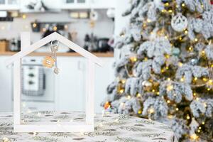 sleutel en klein huis van een klein grootte Aan knus huis met Kerstmis decor Aan tafel van feestelijk wit keuken. geschenk voor nieuw jaar. verzekering, in beweging naar nieuw huis, hypotheek, huur en aankoop echt landgoed foto