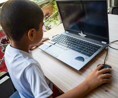 weinig jongen zittend Bij tafel gebruik makend van laptop voor online klasse in rang 1, kind aan het studeren Aan laptop van huis voor afstand aan het leren online opleiding, school- jongen kinderen levensstijl concept foto