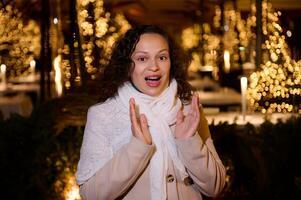 mooi vrouw uitdrukken verbazing staand tegen een stad straat verlichte met Kerstmis slingers Bij nacht tijd foto