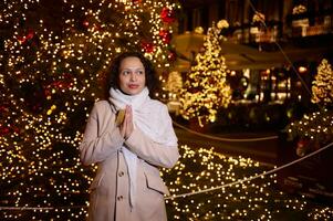 mooi vrouw met tot een kom gevormd handen, glimlacht op zoek terzijde, maakt een gekoesterd wens voor kerstmis, staand tegen een Kerstmis boom met mooi slingers en verlichting in de nacht in Italiaans stad como foto