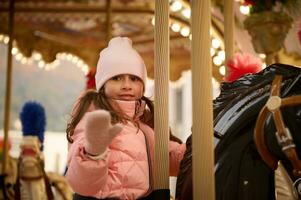 mooi kind meisje rijden een vrolijk Gaan ronde carrousel paard, genieten van gelukkig tijd Bij Kerstmis kermis. foto