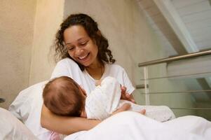 gelukkig liefhebbend moeder glimlacht terwijl verbindt met haar baby, houdt hem in haar armen, zittend Aan de bed in wit slaapkamer foto