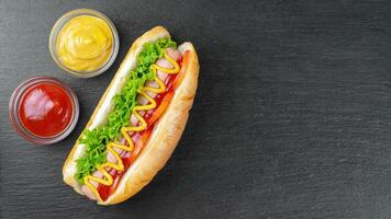 eigengemaakt heet hond met mosterd, ketchup, tomaat en vers salade bladeren Aan zwart leisteen achtergrond foto