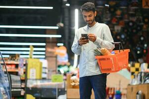 jong Mens gebruik makend van mobiel telefoon terwijl boodschappen doen Bij supermarkt foto