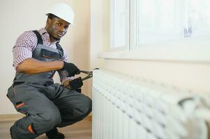 afro klusjesman in overall gebruik makend van gereedschap terwijl installeren of repareren verwarming radiator foto