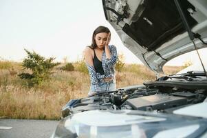 aantrekkelijk slank jong meisje in zomer shorts en overhemd reparaties een gebroken auto. een mooi vrouw staat in de buurt verheven auto kap. foto