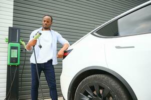 echt Afrikaanse Mens Holding in rekening brengen kabel in Aan hand, staand in de buurt luxe elektrisch auto. foto