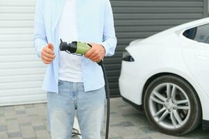 Mens Holding macht opladen kabel voor elektrisch auto in buitenshuis auto park. en hij s gaan naar aansluiten de auto naar de opladen station in de parkeren veel in de buurt de boodschappen doen centrum. foto