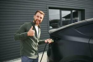 knap gebaard Mens in gewoontjes dragen, staand Bij de opladen station en oplader voor een elektrisch auto. eco elektrisch auto concept foto