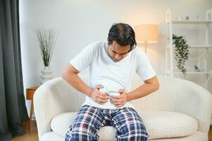 ongelukkig Indisch of Arabisch Mens, zit Aan comfortabel sofa in knus leven kamer, houdt zijn handen Aan zijn maag, grimassen van pijn in zijn maag, lijdt van vergiftiging, spasme, maag problemen. foto