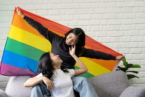gelukkig Aziatisch lgbtq paar zittend Aan sofa en golvend regenboog lgbt trots vlaggen samen in leven kamer Bij huis. concepten van lgbt relaties en lesbienne paren foto