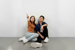 portret van vrolijk jong Aziatisch paar in gewoontjes outfits zittend Aan verdieping en richten Bij kopiëren ruimte foto