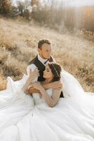 portret van een elegant bruidegom met een bruid Aan een achtergrond van herfst droog gras. de concept van een landelijk bruiloft in de bergen, gelukkig Boheems pasgetrouwden. de bruid en bruidegom zijn aan het liegen Aan de gras foto