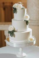 een wit bruiloft taart, versierd met roos bloemen, staat Aan een glas stellage. vers bloemen voor een bruiloft. snoepgoed foto