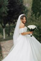bruiloft foto. de bruid in een omvangrijk wit jurk en een lang sluier wandelingen in de tuin langs een steen pad, looks over- haar schouder Bij de camera, Holding een boeket van wit rozen. foto