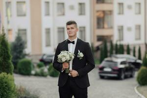 een Mens in een wit shirt, een zwart boog stropdas en een zwart pak houdt een boeket en gaat naar een ontmoeting, achter hem zijn zwart auto's, nieuw gebouwen en bomen. een elegant horloge. Mannen stijl. mode. bedrijf foto