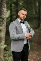 elegant bruidegom bijgestelde zijn kijk maar op zoek in camera lens. luxe duur kijk maar Aan hand. grijs jasje, wit shirt, enkel laarzen gemaakt van vers bloemen. foto details