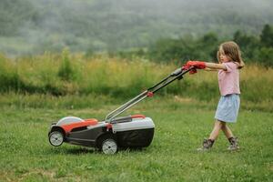 een kind in laarzen in de het formulier van een spel maait gras met een grasmaaier in de werf tegen de achtergrond van bergen en mist, de concept van tuin gereedschap foto