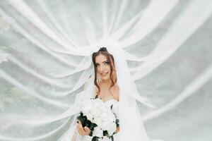 gekruld brunette bruid in een wit jurk, gedekt met een sluier, poses voor de camera met een boeket van rozen. portret van de bruid. mooi bedenken en haar. bruiloft in natuur foto