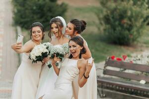 bruiloft foto in natuur. een brunette bruid in een lang wit jurk en haar vrienden in naakt jurken zijn glimlachen en nemen een selfie Aan hun telefoon, Holding hun gypsophila boeketten. jong Dames. emoties