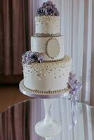 wit drieledig bruiloft taart versierd met Purper bloemen en kralen, staand Aan een spiegel stellage. desserts bruiloft. foto