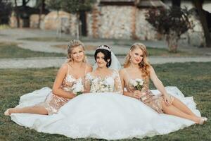 een brunette bruid in een wit elegant jurk met een kroon en haar blond vrienden in goud jurken houding met boeketten terwijl zittend Aan de gras. bruiloft portret in natuur, bruiloft foto in een licht toon.