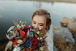 bruid Aan de bank van de rivier, met kaal poten zittend Aan een rots Holding een boeket van divers bloemen, rood rozen en blauw bloemen en een lang wit jurk foto