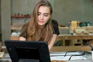 een jong vrouw is opleiding naar worden een timmerman in de werkplaats. ze werken met een laptop computer in een hout werkplaats. vrouw timmerman contact klanten door smartphone. mkb bestellingen, opstarten en klein foto