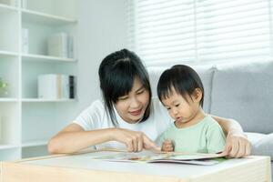 gelukkig Aziatisch moeder kom tot rust en lezen boek met baby tijd samen Bij huis. ouder zitten Aan sofa met dochter en lezing een verhaal. leren ontwikkeling, kinderopvang, lachend, opleiding, verhaal vertellen, oefening. foto