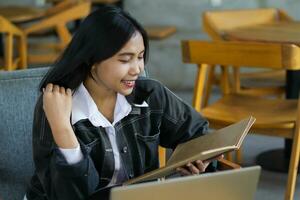 gelukkig Aziatisch vrouw lezing notitieboekje en gebruik makend van laptop in bibliotheek naar aan het studeren foto