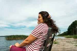 een langharig vrouw is zittend alleen Aan een stoel Aan de kust van een zomer meer, rechttrekken haar haar- foto